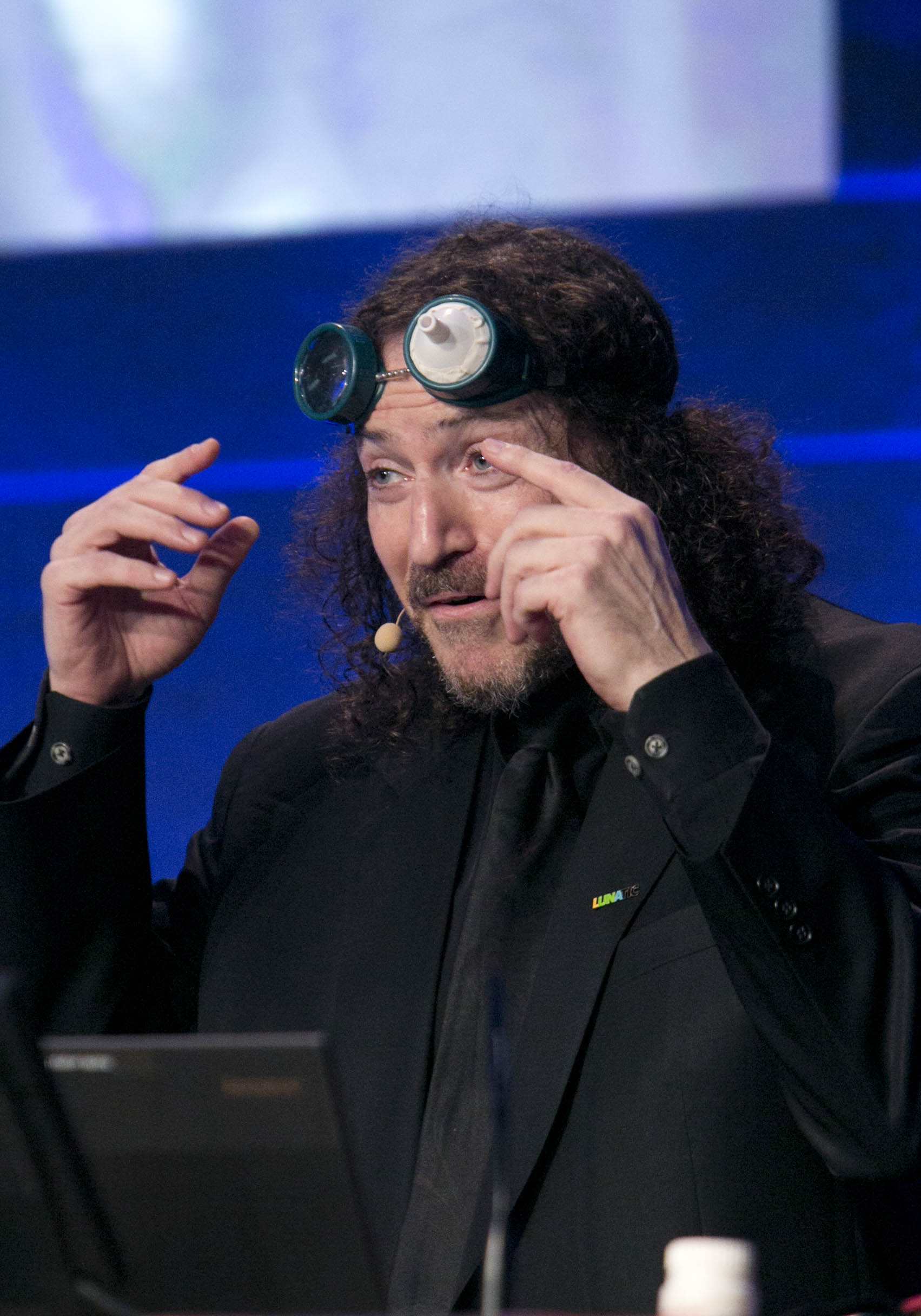 photo of David Berman wearing a microphone, speaking on stage wearing vision impairment simulation goggles on forehead