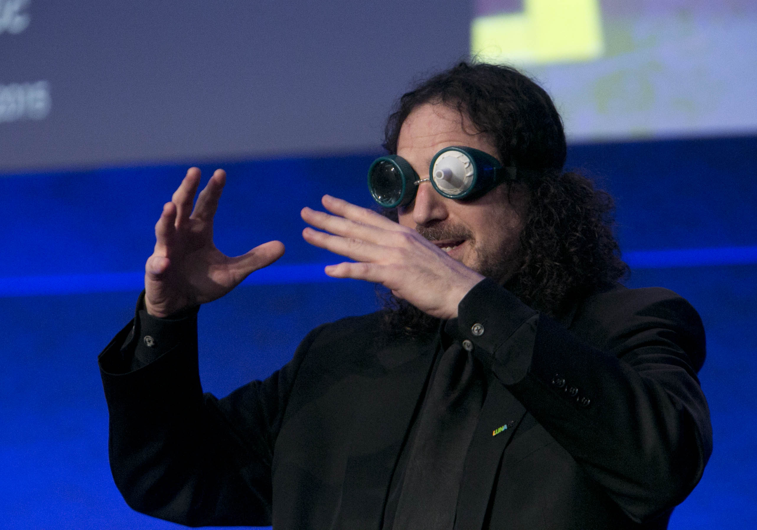photo of David Berman wearing a microphone, speaking on stage wearing vision impairment simulation goggles